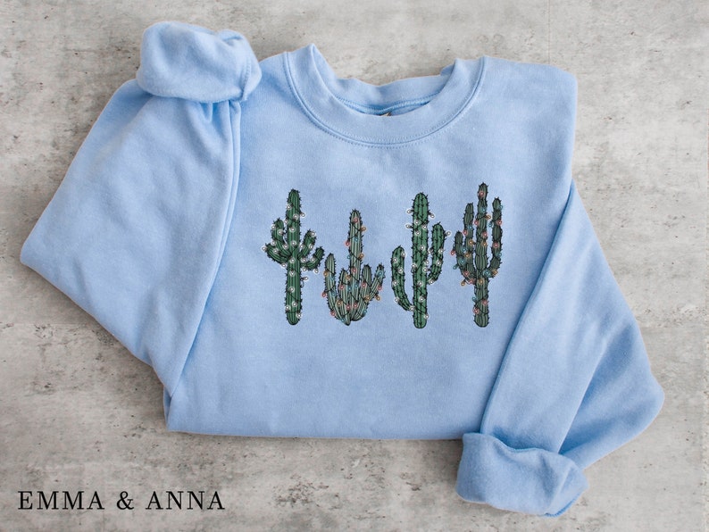 May include: Light blue sweatshirt with a graphic of four cacti with green and brown details and white lights. The cacti are arranged in a row.