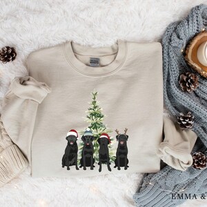 May include: A beige sweatshirt with a Christmas tree and four black Labrador Retrievers wearing Santa hats and reindeer antlers. The dogs are standing in front of the tree.