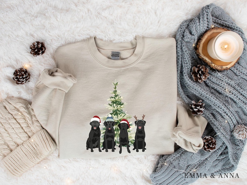 May include: A beige sweatshirt with a Christmas tree and four black Labrador Retrievers wearing Santa hats and reindeer antlers. The dogs are standing in front of the tree.