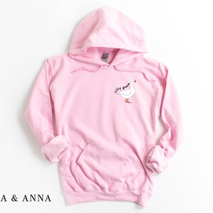 K&ouml;nnte beinhalten: Ein rosa Kapuzenpullover mit einem wei&szlig;en G&auml;nse-Motiv und dem Text "silly goose" auf der Vorderseite.