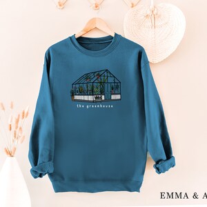 May include: Teal sweatshirt with a greenhouse illustration and the words "the greenhouse." The drawing shows a glass structure filled with plants. The sweatshirt is hanging on a wooden hanger.