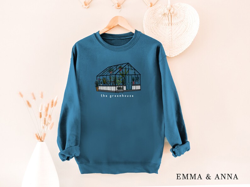 May include: Teal sweatshirt with a greenhouse illustration and the words "the greenhouse." The drawing shows a glass structure filled with plants. The sweatshirt is hanging on a wooden hanger.