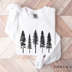 May include: White sweatshirt with a black silhouette of five pine trees printed on the front. The text "EMMA & ANNA" is printed on the bottom right corner of the sweatshirt.