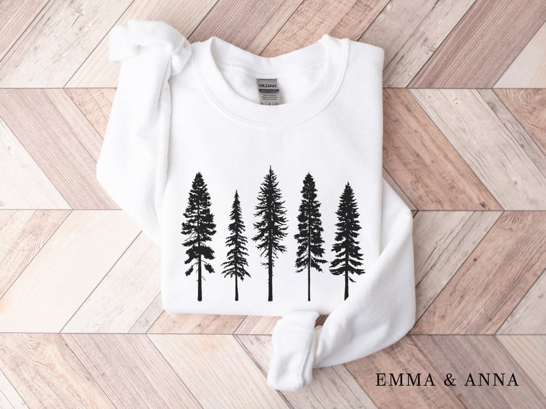 May include: White sweatshirt with a black silhouette of five pine trees printed on the front. The text "EMMA & ANNA" is printed on the bottom right corner of the sweatshirt.