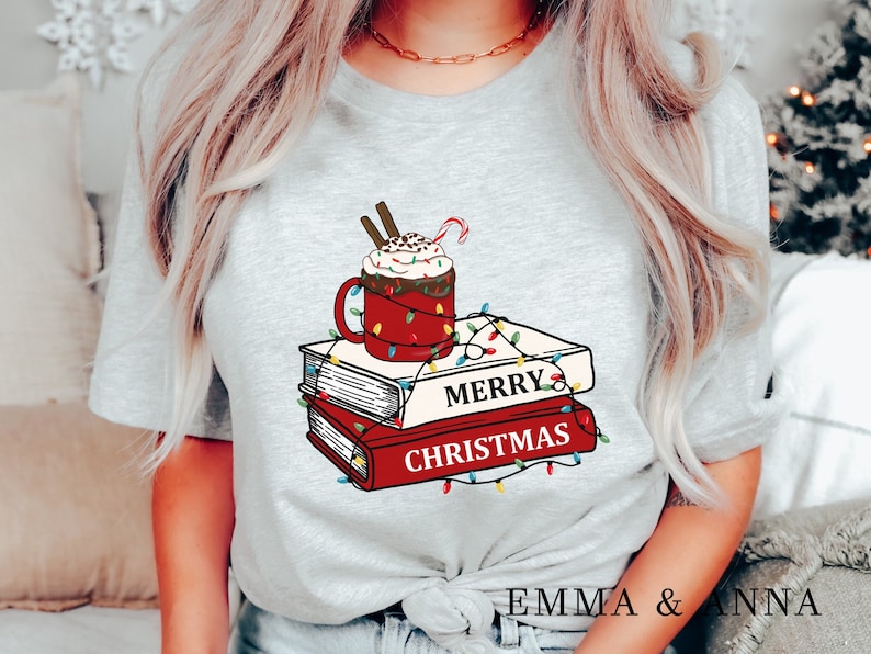 May include: A gray t-shirt with a red mug of hot chocolate topped with whipped cream and a candy cane. The mug is surrounded by colorful Christmas lights and a stack of books with the text "Merry Christmas".