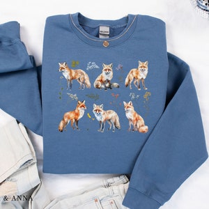 May include: A blue sweatshirt with a print of six foxes in different poses. The foxes are surrounded by small floral designs. The sweatshirt has a crew neck and long sleeves.