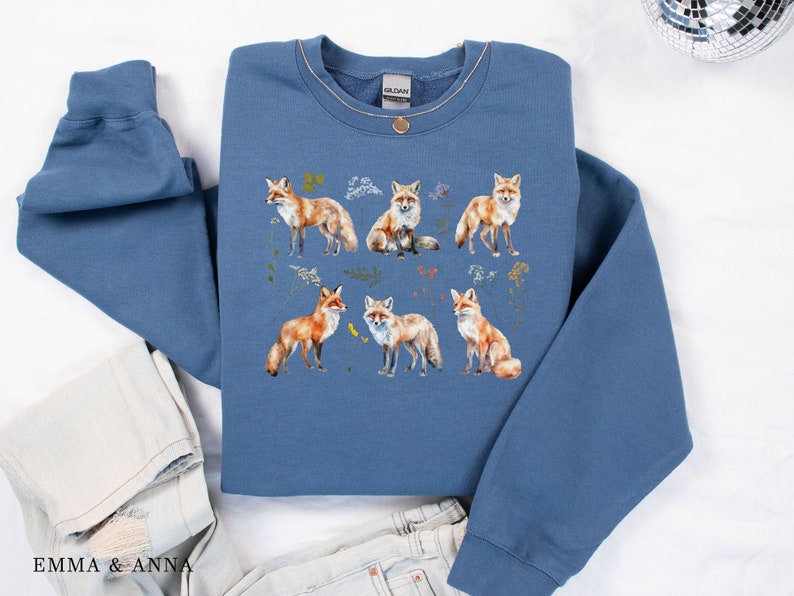 May include: A blue sweatshirt with a print of six foxes in different poses. The foxes are surrounded by small floral designs. The sweatshirt has a crew neck and long sleeves.
