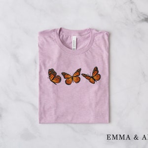 Może przedstawiać: Jasnofioletowa koszulka z trzema pomarańczowo-czarnymi motywami motyli, wyszytymi z przodu. Nazwa marki "EMMA & ANNA" jest wydrukowana na dole po prawej stronie.
