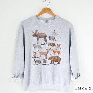 May include: A gray sweatshirt with a graphic print of various woodland animals, including a moose, wolf, deer, fox, bear, raccoon, squirrel, chipmunk, badger, hedgehog, and hare. The animals are illustrated in a realistic style with brown, black, white, and gray colors.