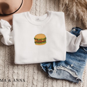 May include: White sweatshirt with a cartoon illustration of a hamburger on the front. The sweatshirt is folded and laid on a beige knitted surface with a pair of blue denim shorts and a brown hat.