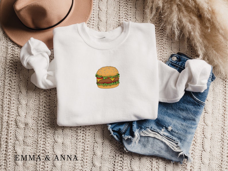 May include: White sweatshirt with a cartoon illustration of a hamburger on the front. The sweatshirt is folded and laid on a beige knitted surface with a pair of blue denim shorts and a brown hat.
