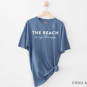 Pu&ograve; includere: Una maglietta blu con la scritta "THE BEACH is my therapy" in corsivo bianco. La maglietta &egrave; appesa a una gruccia di legno su sfondo bianco. Il fondo della maglietta &egrave; annodato.