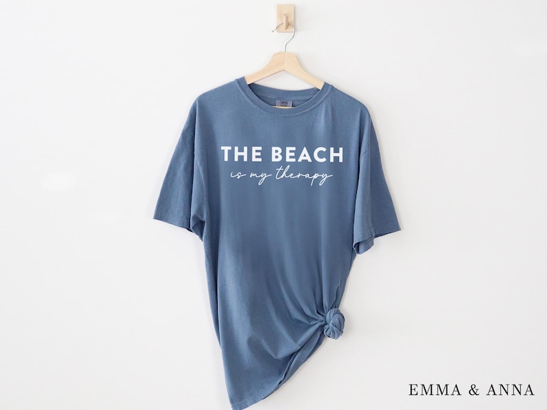 Pu&ograve; includere: Una maglietta blu con la scritta "THE BEACH is my therapy" in corsivo bianco. La maglietta &egrave; appesa a una gruccia di legno su sfondo bianco. Il fondo della maglietta &egrave; annodato.