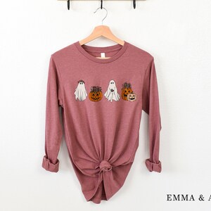 May include: A long-sleeved mauve t-shirt with a graphic of ghosts and pumpkins wearing floral crowns. The shirt is tied in a knot at the bottom.