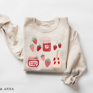 May include: A beige sweatshirt with a strawberry jam theme. The sweatshirt features a red and white jar with the words "Strawberry Jam" and several red strawberries with green leaves.