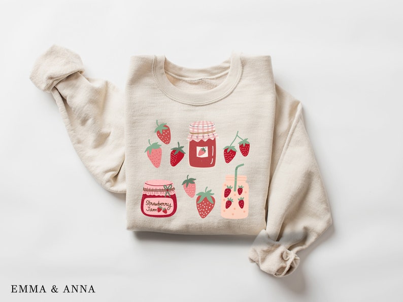 May include: A beige sweatshirt with a strawberry jam theme. The sweatshirt features a red and white jar with the words "Strawberry Jam" and several red strawberries with green leaves.