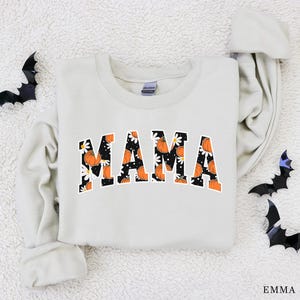Op de afbeelding: Cr&egrave;mekleurige sweatshirt met het woord "MAMA" in oranje letters gevuld met een pompoen- en madeliefpatroon. Zwarte vleermuisdecoraties zijn rond de sweatshirt geplaatst. De sweatshirt is een herfstthema-artikel.