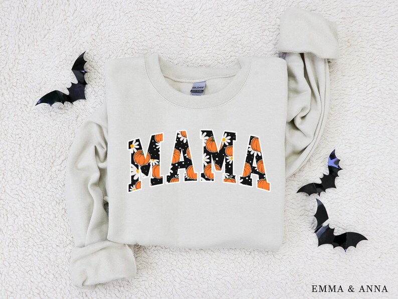 Op de afbeelding: Cr&egrave;mekleurige sweatshirt met het woord "MAMA" in oranje letters gevuld met een pompoen- en madeliefpatroon. Zwarte vleermuisdecoraties zijn rond de sweatshirt geplaatst. De sweatshirt is een herfstthema-artikel.