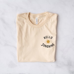May include: A light beige t-shirt with a black graphic that says "Hello Sunshine" with a yellow sun graphic.
