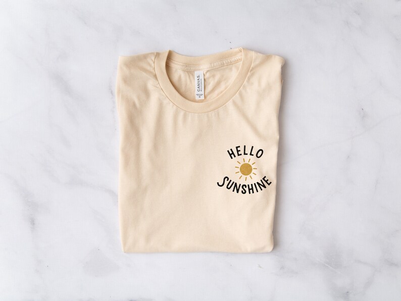 May include: A light beige t-shirt with a black graphic that says "Hello Sunshine" with a yellow sun graphic.