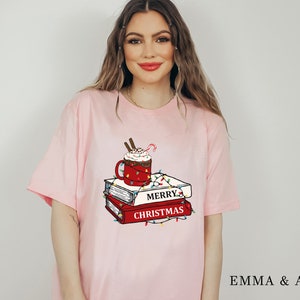May include: Pink t-shirt with a graphic of a red mug with whipped cream and a candy cane, a stack of books, and Christmas lights. The text on the books reads "Merry Christmas".