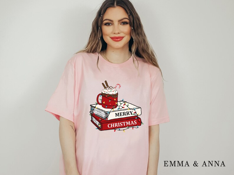 May include: Pink t-shirt with a graphic of a red mug with whipped cream and a candy cane, a stack of books, and Christmas lights. The text on the books reads "Merry Christmas".