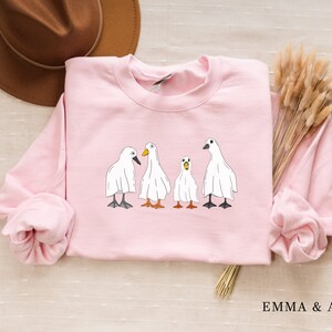 May include: A pink sweatshirt with a graphic of four white ducks wearing ghost costumes. The ducks are lined up in a row, with the one in the middle looking slightly surprised.