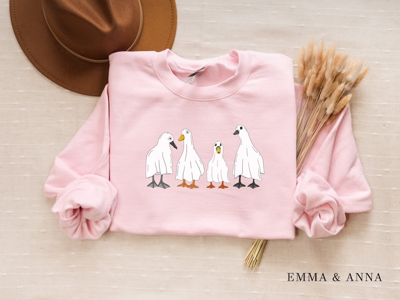 May include: A pink sweatshirt with a graphic of four white ducks wearing ghost costumes. The ducks are lined up in a row, with the one in the middle looking slightly surprised.