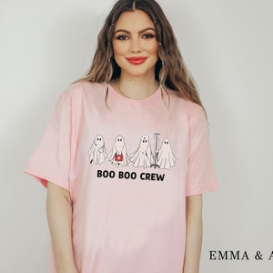 Puede incluir: Camiseta rosa con un gr&aacute;fico de cuatro fantasmas vestidos de enfermeras. El texto "BOO BOO CREW" est&aacute; impreso debajo de los fantasmas.