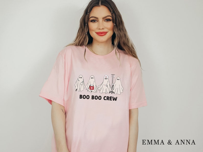 Puede incluir: Camiseta rosa con un gr&aacute;fico de cuatro fantasmas vestidos de enfermeras. El texto "BOO BOO CREW" est&aacute; impreso debajo de los fantasmas.