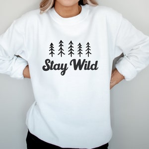 K&ouml;nnte beinhalten: Wei&szlig;er Pullover mit einer schwarzen Grafik einer Reihe von Kiefern und dem Text "Stay Wild".