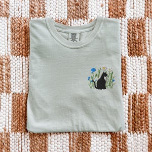 May include: A light green t-shirt with a crew neck. The shirt features an embroidered design of a black cat sitting in a field of white daisies, blue flowers, and green grass. The shirt is folded neatly on a patterned surface.