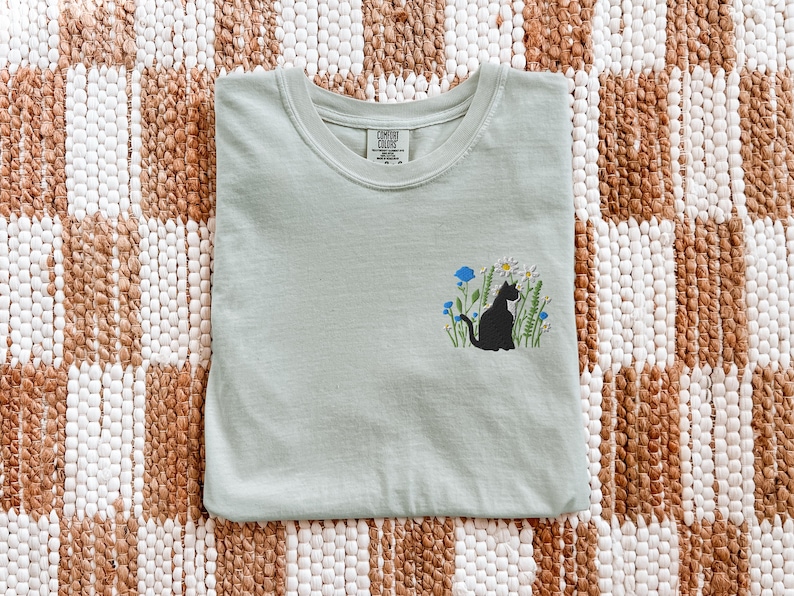 May include: A light green t-shirt with a crew neck. The shirt features an embroidered design of a black cat sitting in a field of white daisies, blue flowers, and green grass. The shirt is folded neatly on a patterned surface.