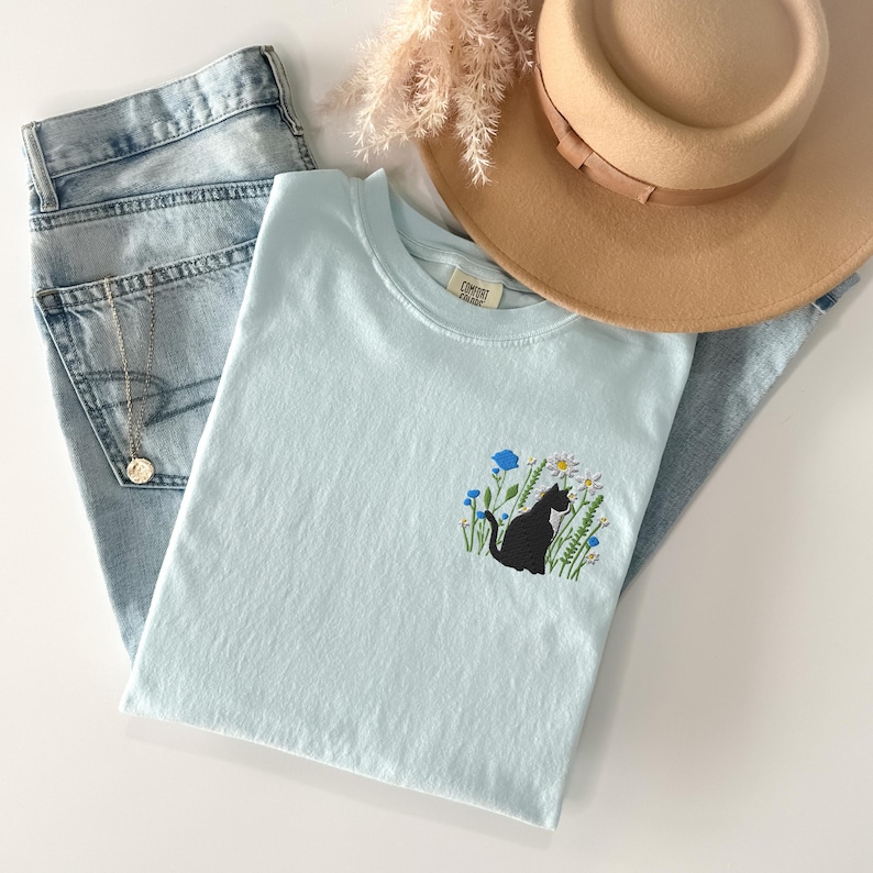 May include: A light blue t-shirt with a black cat embroidered on the front, surrounded by flowers. The shirt is folded next to a pair of light blue jeans, a tan hat, and a gold necklace.