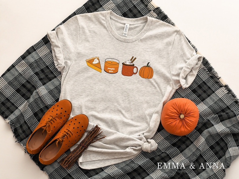 May include: A gray t-shirt with a pumpkin spice latte, pumpkin pie, and a pumpkin graphic. The shirt is laying on a black and white plaid blanket with two brown shoes and a brown pumpkin.