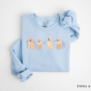 May include: Light blue sweatshirt with a graphic of four golden retrievers sitting in a row with flowers and greenery. The sweatshirt has a crew neck and long sleeves.