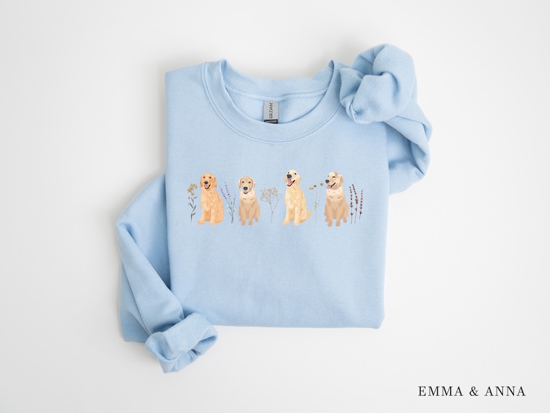 May include: Light blue sweatshirt with a graphic of four golden retrievers sitting in a row with flowers and greenery. The sweatshirt has a crew neck and long sleeves.