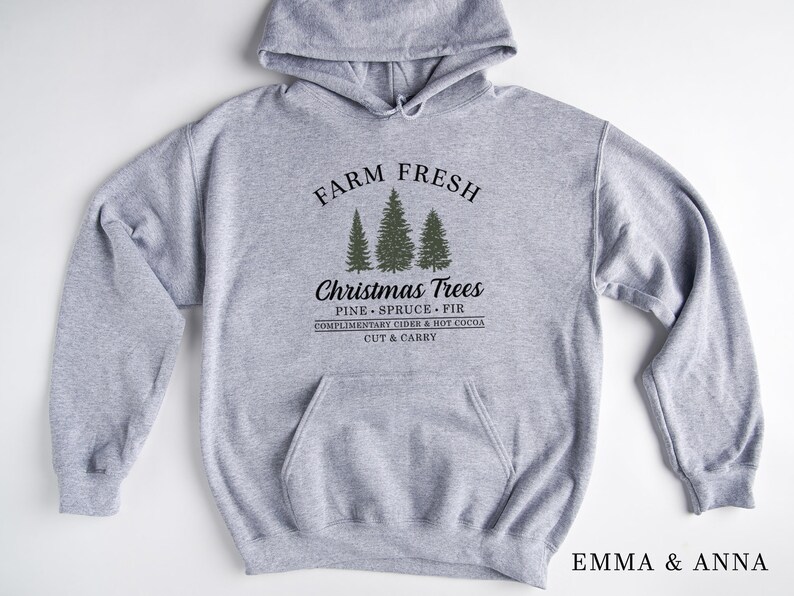 Op de afbeelding: Grijs hooded sweatshirt met de tekst "FARM FRESH" boven een afbeelding van drie naaldbomen en de tekst "Christmas Trees PINE SPRUCE FIR COMPLIMENTARY CIDER & HOT COCOA CUT & CARRY" onder de bomen.