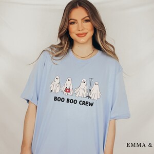 Puede incluir: Una camiseta azul claro con un gr&aacute;fico de cuatro fantasmas vestidos como profesionales m&eacute;dicos. El texto "BOO BOO CREW" est&aacute; impreso debajo de los fantasmas.