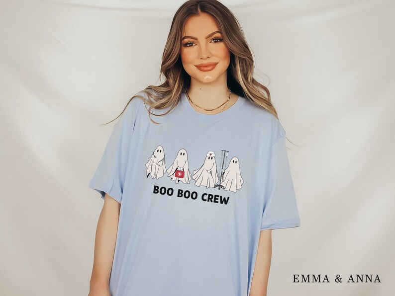 Puede incluir: Una camiseta azul claro con un gr&aacute;fico de cuatro fantasmas vestidos como profesionales m&eacute;dicos. El texto "BOO BOO CREW" est&aacute; impreso debajo de los fantasmas.