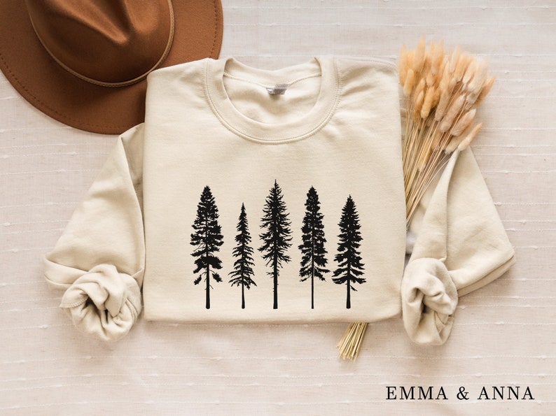 May include: A cream-colored sweatshirt with a black silhouette of five pine trees printed on the front. The sweatshirt has long sleeves and a crew neck.