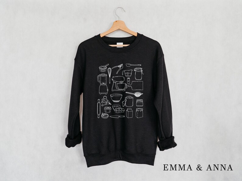 May include: Black sweatshirt with a white graphic design of various kitchen tools and baking supplies. The design includes a stand mixer, measuring cups, whisks, and more.