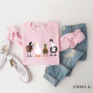 Peut inclure: Sweat-shirt rose avec un motif de quatre canards align&eacute;s. Les canards sont de couleurs et de races diff&eacute;rentes. Le sweat-shirt est pos&eacute; &agrave; plat sur une surface blanche avec une paire de baskets blanches et un jean bleu.