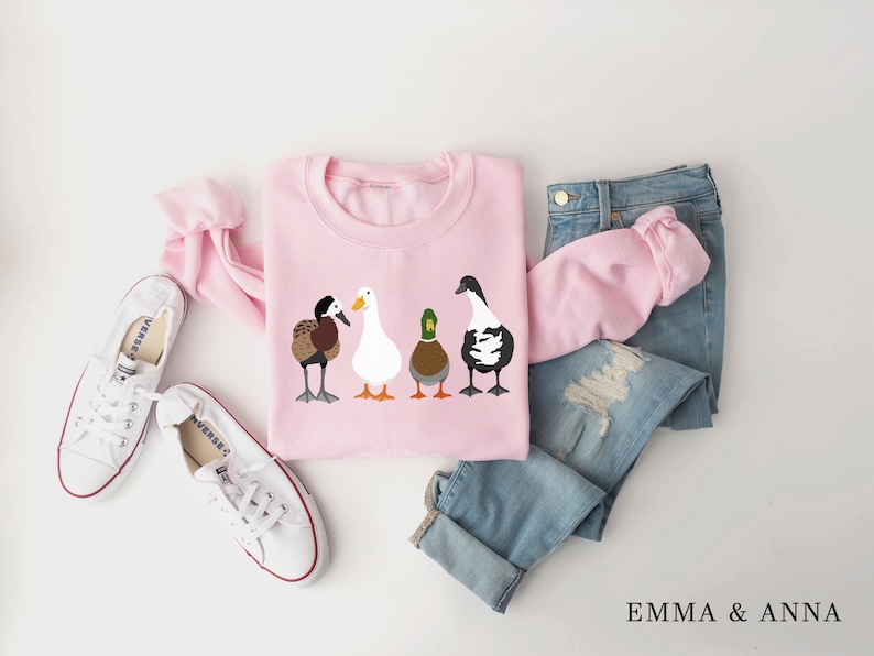 Peut inclure: Sweat-shirt rose avec un motif de quatre canards align&eacute;s. Les canards sont de couleurs et de races diff&eacute;rentes. Le sweat-shirt est pos&eacute; &agrave; plat sur une surface blanche avec une paire de baskets blanches et un jean bleu.