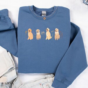 May include: A blue sweatshirt with a graphic of four golden retriever dogs in a row, each with a different expression. The dogs are surrounded by small flowers and leaves.