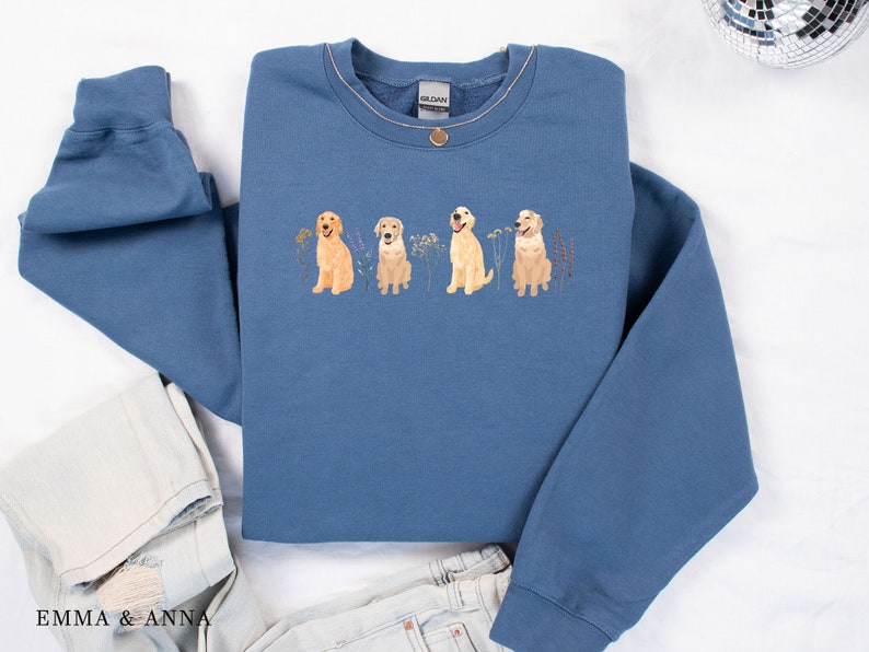 May include: A blue sweatshirt with a graphic of four golden retriever dogs in a row, each with a different expression. The dogs are surrounded by small flowers and leaves.