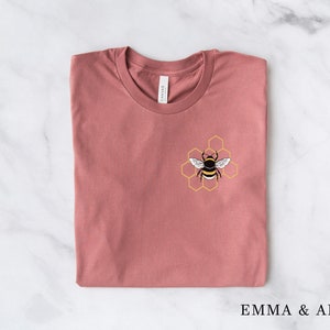 May include: A folded mauve t-shirt with a bee and honeycomb design on the front. The design is in yellow and black.