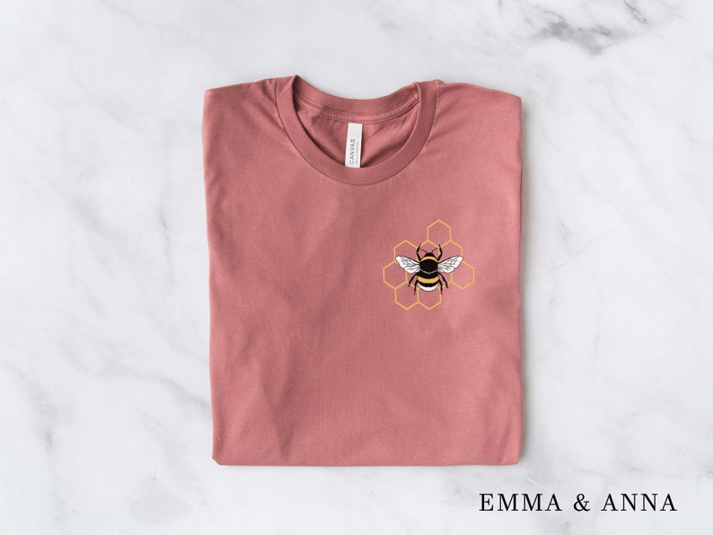 May include: A folded mauve t-shirt with a bee and honeycomb design on the front. The design is in yellow and black.