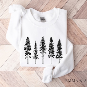 Puede incluir: Sudadera blanca con una silueta negra de cinco pinos impresa en la parte delantera. El texto "EMMA & ANNA" est&aacute; impreso en la esquina inferior derecha de la sudadera.