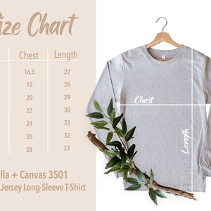 Puede incluir: Una camiseta de manga larga unisex de punto de jersey gris claro cuelga de una percha de madera. La imagen incluye una tabla de tallas con medidas de pecho y largo en cent&iacute;metros. La tabla enumera las tallas XS a 3XL. Una ramita de vegetaci&oacute;n a&ntilde;ade un toque natural.